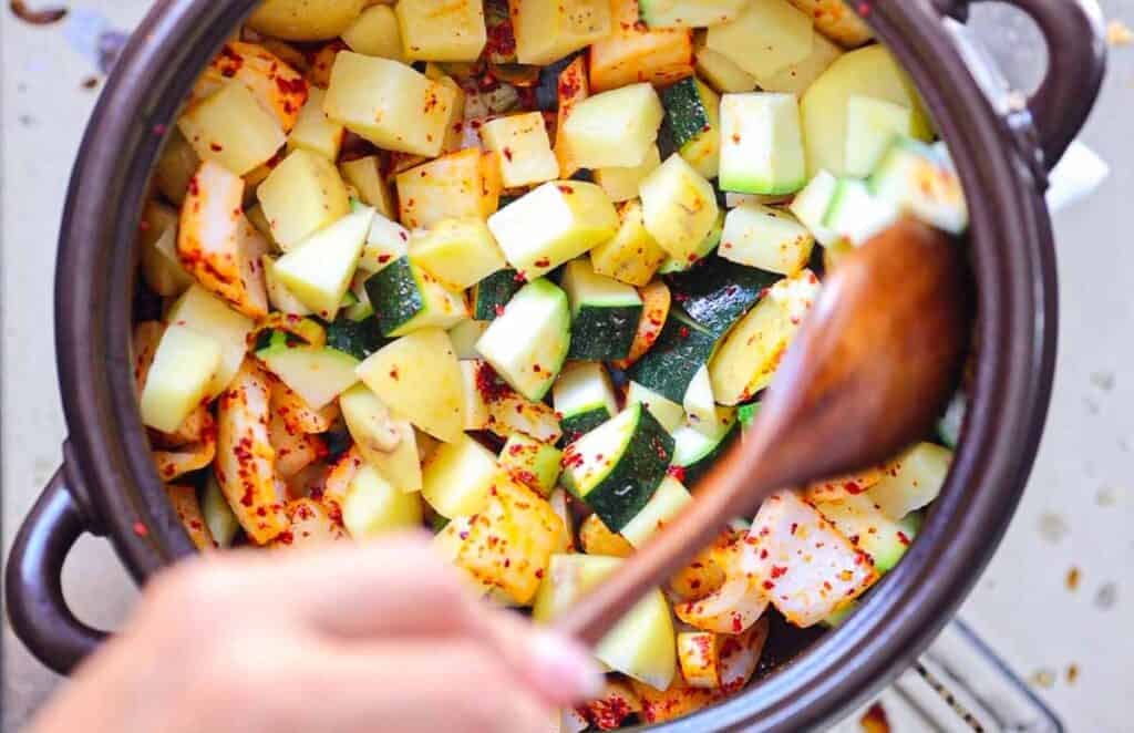 A pot filled with chopped potatoes and zucchini seasoned with red pepper flakes. A hand is stirring the mixture with a wooden spoon. The vegetables are vibrant, with green and yellow hues, contrasting with the red spices.